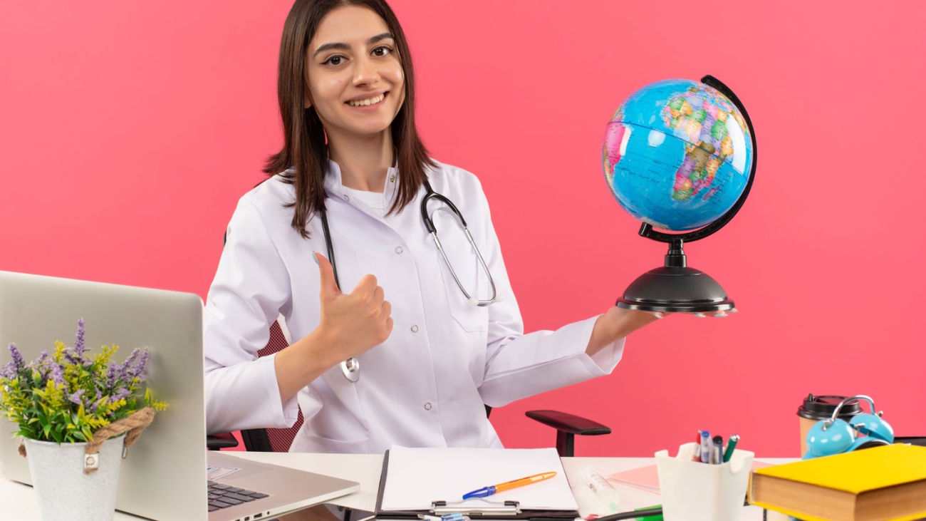 young-female-doctor-white-coat-with-stethoscope-around-her-neck-holding-globe-looking-front-with-smile-face-showing-thumbs-up-sitting-table-with-laptop-pink-wall