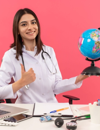 young-female-doctor-white-coat-with-stethoscope-around-her-neck-holding-globe-looking-front-with-smile-face-showing-thumbs-up-sitting-table-with-laptop-pink-wall